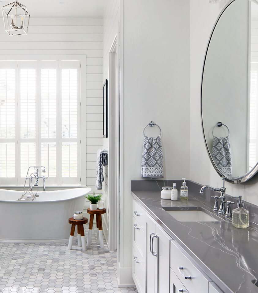 Bright white bathroom with dark gray marble countertops, gray and cream hexagon tile, and two decorative wooden stools aside bathtub
